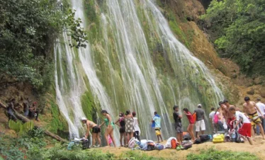 A group in swimwear gathers at the base of a lush, moss-covered waterfall, surrounded by vibrant vegetation—some stand while others relax near their bags and towels, savoring a day in nature on one of many unforgettable Punta Cana excursions.