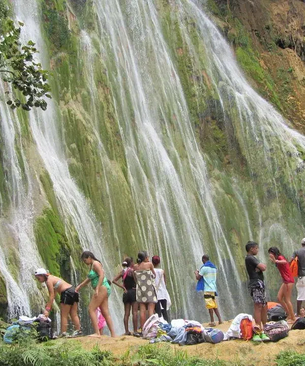 A group in swimwear gathers at the base of a lush, moss-covered waterfall, surrounded by vibrant vegetation—some stand while others relax near their bags and towels, savoring a day in nature on one of many unforgettable Punta Cana excursions.