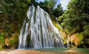 A tall, wide waterfall cascades over mossy rocks into a turquoise pool, surrounded by lush green foliage under a sunny sky—a tranquil, tropical scene you can discover on unforgettable Punta Cana tours.
