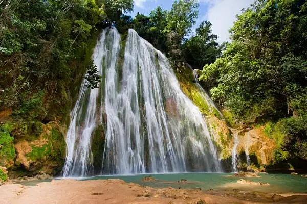 A tall, wide waterfall cascades over mossy rocks into a turquoise pool, surrounded by lush green foliage under a sunny sky—a tranquil, tropical scene you can discover on unforgettable Punta Cana tours.