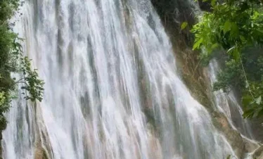 A large group of people stands at the base of a wide, cascading waterfall surrounded by green trees and foliage—a highlight of many Punta Cana excursions. Mist rises as water flows down the rocky cliff, creating a lively natural setting.