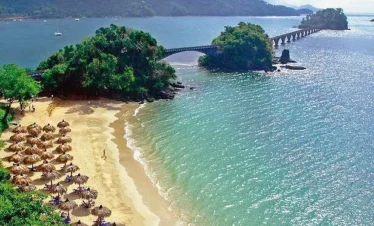 A sandy beach with rows of thatched umbrellas faces turquoise water. Lush green trees cover a small island linked by a long, arched bridge—a dreamy scene perfect for tours in Punta Cana, with hills and boats under a mostly clear sky.