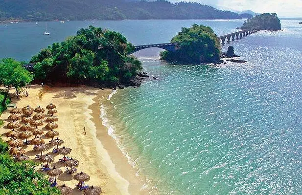 A sandy beach with rows of thatched umbrellas faces turquoise water. Lush green trees cover a small island linked by a long, arched bridge—a dreamy scene perfect for tours in Punta Cana, with hills and boats under a mostly clear sky.