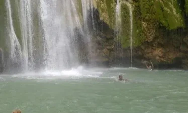 A tall waterfall cascades down a moss-covered cliff into a turquoise pool. Two people swim below, surrounded by rocks and greenery—a tranquil outdoor setting perfect for those seeking adventure with Punta Cana tours.