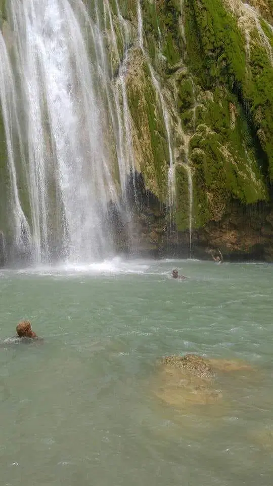 A tall waterfall cascades down a moss-covered cliff into a turquoise pool. Two people swim below, surrounded by rocks and greenery—a tranquil outdoor setting perfect for those seeking adventure with Punta Cana tours.