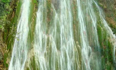 Tall, multi-stream waterfall cascades down a moss-covered rocky cliff into a turquoise pool below, surrounded by lush green foliage and sunlight—an idyllic spot often featured on tours in Punta Cana.