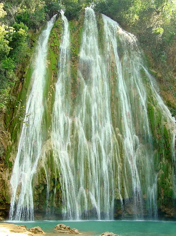 Tall, multi-stream waterfall cascades down a moss-covered rocky cliff into a turquoise pool below, surrounded by lush green foliage and sunlight—an idyllic spot often featured on tours in Punta Cana.