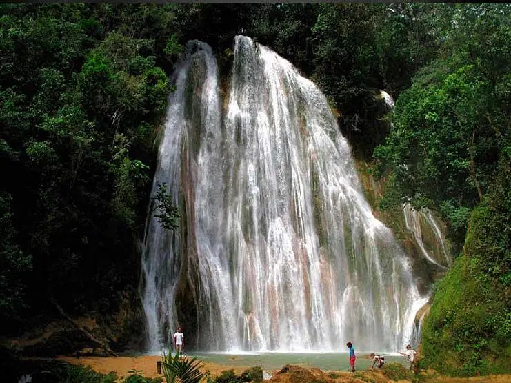A wide, powerful waterfall cascades down a lush green cliff into a pool below. Four people stand near the water’s edge, some wading in—an ideal scene to discover on Punta Cana excursions amid dense forest and vibrant greenery under natural daylight.
