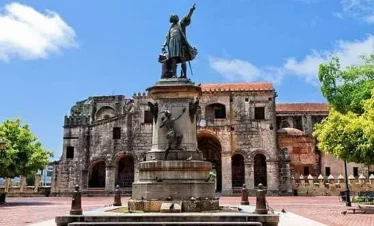 A stone statue of Christopher Columbus stands on a pedestal in a plaza, with the Cathedral of Santa María la Menor behind it—an iconic sight often included in Punta Cana tours for those seeking history beyond their Punta Cana trip.