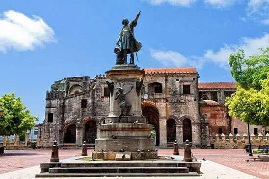 A stone statue of Christopher Columbus stands on a pedestal in a plaza, with the Cathedral of Santa María la Menor behind it—an iconic sight often included in Punta Cana tours for those seeking history beyond their Punta Cana trip.