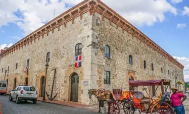 A large, historic stone building with arched doors and small windows is shown. The Dominican Republic flag hangs on the wall. Nearby, a horse-drawn carriage and parked cars await—perfect sights to experience on Punta Cana tours under a blue sky.