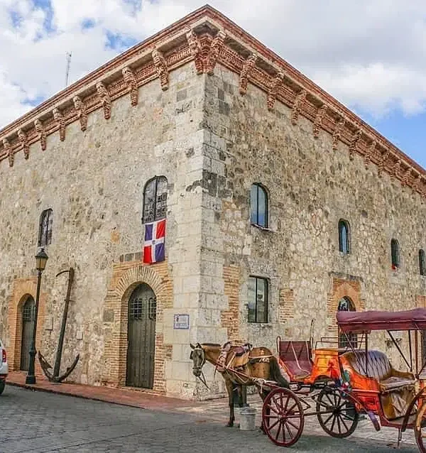 A large, historic stone building with arched doors and small windows is shown. The Dominican Republic flag hangs on the wall. Nearby, a horse-drawn carriage and parked cars await—perfect sights to experience on Punta Cana tours under a blue sky.