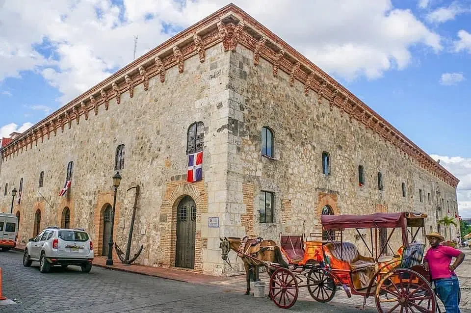 A large, historic stone building with arched doors and small windows is shown. The Dominican Republic flag hangs on the wall. Nearby, a horse-drawn carriage and parked cars await—perfect sights to experience on Punta Cana tours under a blue sky.
