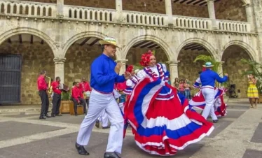 A group of dancers in traditional, colorful costumes—blue, red, and white—perform outside a historic stone building as part of Punta Cana tours. Musicians play in the background while the dancers captivate visitors with vibrant energy.