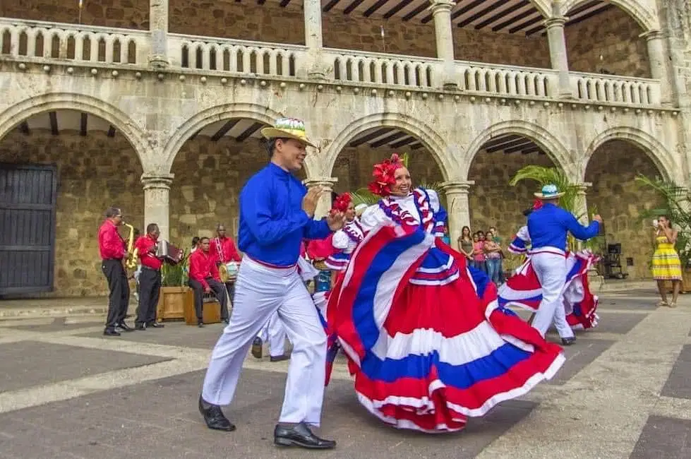 A group of dancers in traditional, colorful costumes—blue, red, and white—perform outside a historic stone building as part of Punta Cana tours. Musicians play in the background while the dancers captivate visitors with vibrant energy.