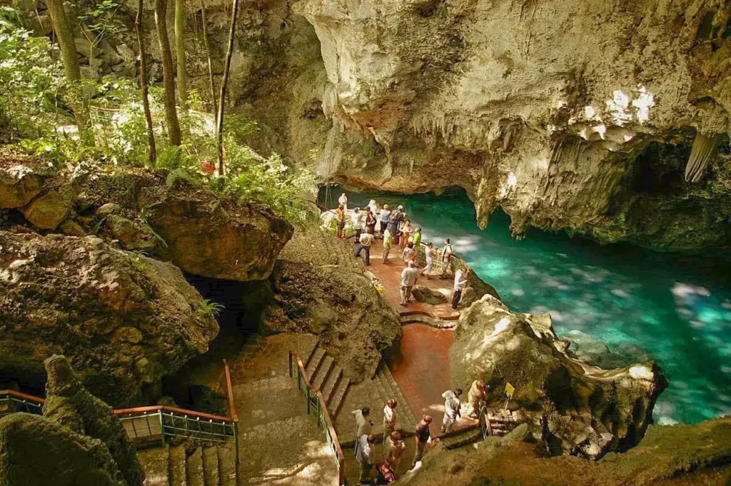 A group of people gather by a turquoise underground pool surrounded by rugged limestone formations and lush greenery. Stone stairs lead down to the water—an enchanting spot often visited on Punta Cana tours for its natural beauty.