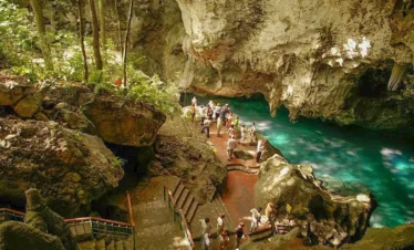 A group of people gather by a turquoise underground pool surrounded by rugged limestone formations and lush greenery. Stone stairs lead down to the water—an enchanting spot often visited on Punta Cana tours for its natural beauty.