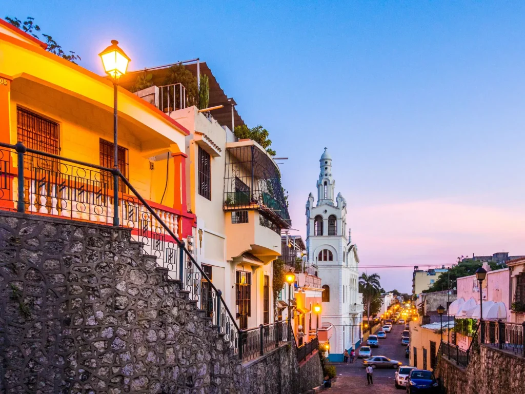 Colorful houses line a sloping cobblestone street at sunset, with warm streetlights glowing. Reminiscent of charming tours in Punta Cana, a tall white church rises in the background; greenery decorates balconies as the sky fades from blue to pink.