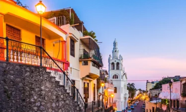Colorful houses line a sloping cobblestone street at sunset, with warm streetlights glowing. Reminiscent of charming tours in Punta Cana, a tall white church rises in the background; greenery decorates balconies as the sky fades from blue to pink.