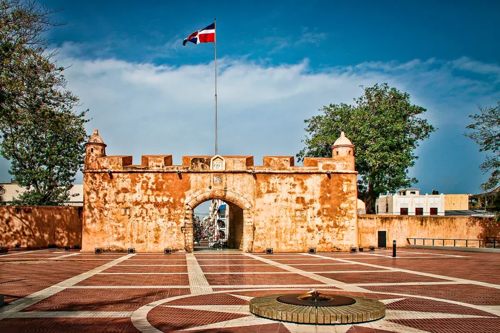 Historic fortified gate with an arched entrance stands on a brick plaza, surrounded by low walls and turrets. A Dominican Republic flag flies overhead—a must-see stop on many Punta Cana tours. Trees and modern buildings rise under a blue sky.