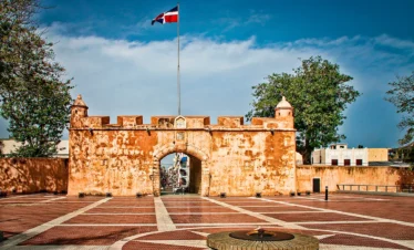 Historic fortified gate with an arched entrance stands on a brick plaza, surrounded by low walls and turrets. A Dominican Republic flag flies overhead—a must-see stop on many Punta Cana tours. Trees and modern buildings rise under a blue sky.