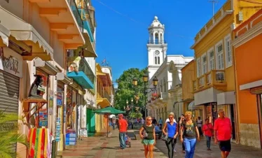 A lively street scene in a historic town with colorful buildings, a white clock tower in the background, and people walking along a sunny pedestrian pathway—perfect for those seeking vibrant sights on Punta Cana excursions.