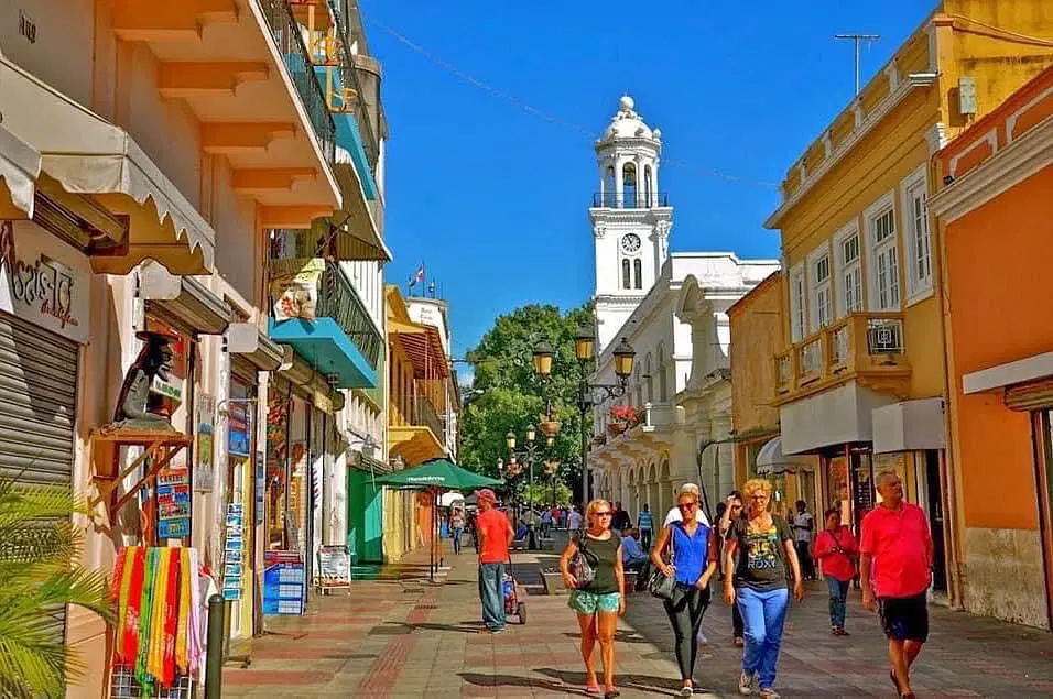 A lively street scene in a historic town with colorful buildings, a white clock tower in the background, and people walking along a sunny pedestrian pathway—perfect for those seeking vibrant sights on Punta Cana excursions.