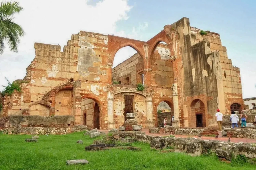 Ruins of a large, old stone and brick building with arched windows and doorways, weathered walls, and missing sections. Grass and plants grow around the site, which is explored by a few people on Punta Cana excursions under a partly cloudy sky.
