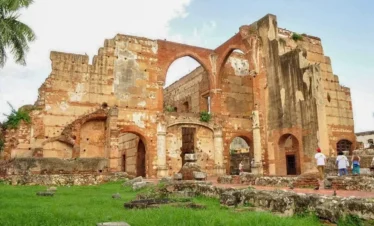 Ruins of a large, old stone and brick building with arched windows and doorways, weathered walls, and missing sections. Grass and plants grow around the site, which is explored by a few people on Punta Cana excursions under a partly cloudy sky.