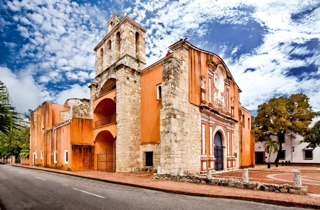 A historic stone church with orange stucco walls and an arched entrance stands on a quiet street—a highlight often featured on Punta Cana tours. It boasts colonial architecture, a bell tower, palm trees, and a brick walkway under blue skies.