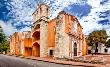 A historic stone church with orange stucco walls and an arched entrance stands on a quiet street—a highlight often featured on Punta Cana tours. It boasts colonial architecture, a bell tower, palm trees, and a brick walkway under blue skies.