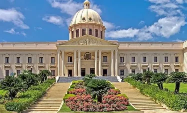 A grand neoclassical building with a large central dome, tall columns, and ornate details is a highlight on many Punta Cana excursions. Manicured gardens line the wide staircase leading to the entrance under a bright blue sky with scattered clouds.
