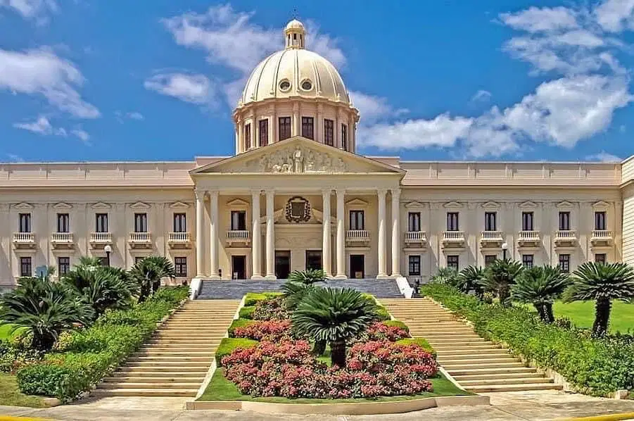 A grand neoclassical building with a large central dome, tall columns, and ornate details is a highlight on many Punta Cana excursions. Manicured gardens line the wide staircase leading to the entrance under a bright blue sky with scattered clouds.