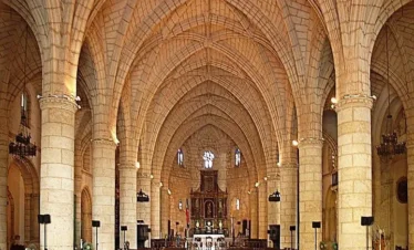 Interior of a grand stone church with vaulted ceilings and ribbed arches, often admired during tours in Punta Cana. Rows of wooden pews lead to an ornate altar, while stained glass windows illuminate the intricate stonework.