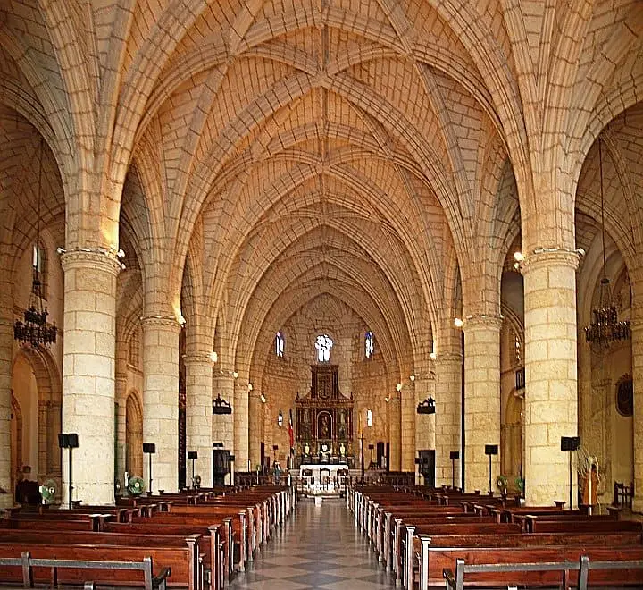 Interior of a grand stone church with vaulted ceilings and ribbed arches, often admired during tours in Punta Cana. Rows of wooden pews lead to an ornate altar, while stained glass windows illuminate the intricate stonework.