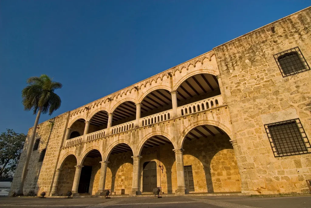 A historic stone building with arched colonnades on both floors, iron-barred windows, and a palm tree on the left—an architectural gem often admired during Punta Cana excursions under the sunlit blue sky.
