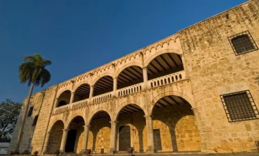 A historic stone building with arched colonnades on both floors, iron-barred windows, and a palm tree on the left—an architectural gem often admired during Punta Cana excursions under the sunlit blue sky.