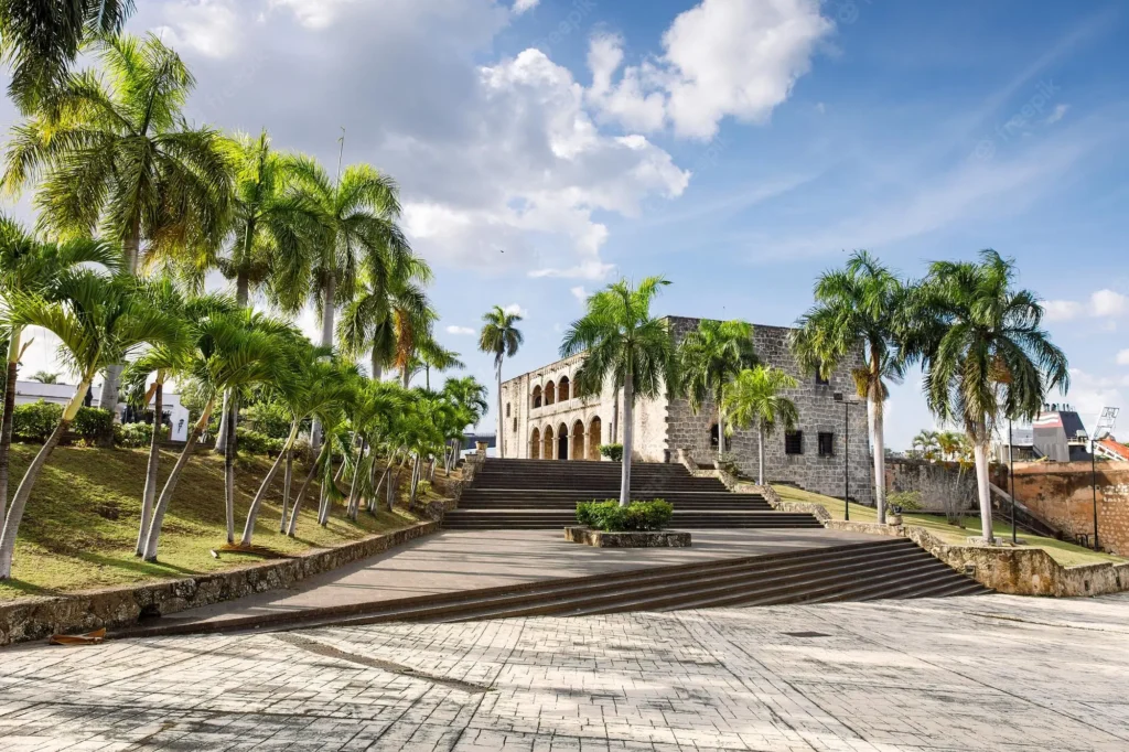 A wide stone staircase lined with palm trees leads to a historic colonial building with arched windows—a perfect spot to discover on Punta Cana excursions beneath the open, sunlit sky, with lush greenery flanking both sides of the steps.
