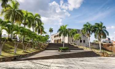 A wide stone staircase lined with palm trees leads to a historic colonial building with arched windows—a perfect spot to discover on Punta Cana excursions beneath the open, sunlit sky, with lush greenery flanking both sides of the steps.