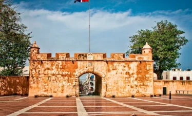 A historic stone fort gate with warm, orange walls stands under a clear blue sky. A Dominican Republic flag flies above—just one of the sights you can explore during unforgettable tours in Punta Cana. The plaza features a monument and flame, flanked by trees.