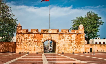 A historic stone fort gate with warm, orange walls stands under a clear blue sky. A Dominican Republic flag flies above—just one of the sights you can explore during unforgettable tours in Punta Cana. The plaza features a monument and flame, flanked by trees.
