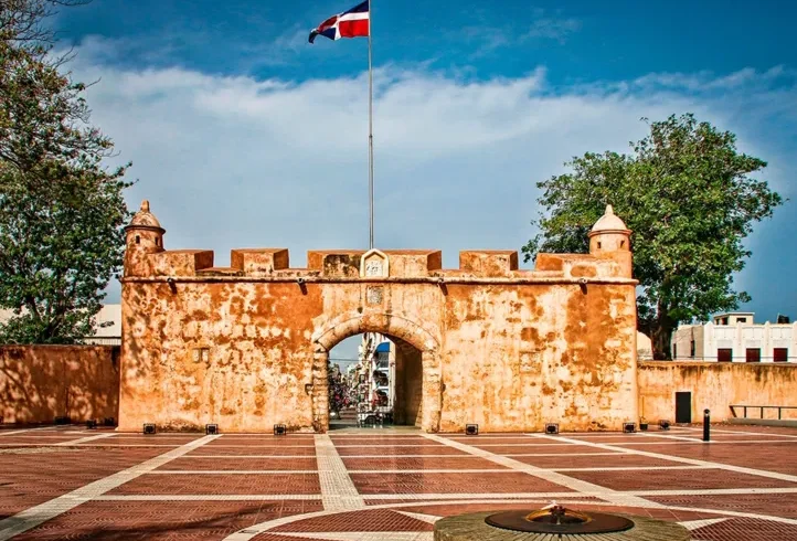 A historic stone fort gate with warm, orange walls stands under a clear blue sky. A Dominican Republic flag flies above—just one of the sights you can explore during unforgettable tours in Punta Cana. The plaza features a monument and flame, flanked by trees.