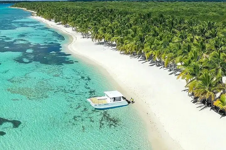 A small boat sits on clear turquoise water at the edge of a white sandy beach in Punta Cana, perfect for tours en punta cana. The shoreline curves gently past lush green palm trees, with coral patches in the sea and dense forest beyond.