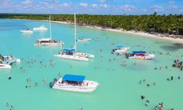 A tropical beach scene in Punta Cana with turquoise water, white boats and catamarans, and people swimming and relaxing. Lush green palm trees and white sand line the shore—perfect for unforgettable tours en Punta Cana under a blue sky with scattered clouds.