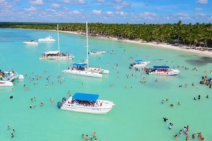 A tropical beach scene in Punta Cana with turquoise water, white boats and catamarans, and people swimming and relaxing. Lush green palm trees and white sand line the shore—perfect for unforgettable tours en Punta Cana under a blue sky with scattered clouds.