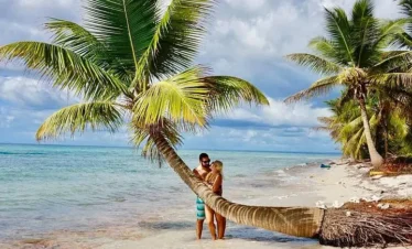 A couple embraces on a sandy tropical beach in Punta Cana, standing by a large leaning palm tree at the shore—an ideal scene for tours in Punta Cana. The calm turquoise ocean and lush greenery create a perfect getaway.