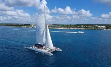 A large white sailing catamaran with raised sails glides on clear blue water, carrying many passengers enjoying one of the best tours in Punta Cana. Another smaller motorboat is in the background near the shoreline under a sky of puffy clouds.