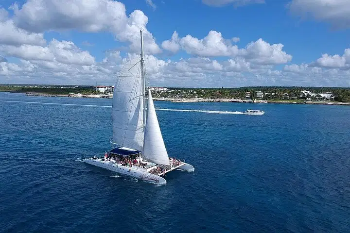 A large white sailing catamaran with raised sails glides on clear blue water, carrying many passengers enjoying one of the best tours in Punta Cana. Another smaller motorboat is in the background near the shoreline under a sky of puffy clouds.