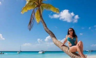 A woman in a teal bikini and sunglasses sits on a curved palm tree overhanging a sandy beach. The turquoise ocean, boats, and bright sky create the perfect scene to experience with the best tours Punta Cana has to offer.
