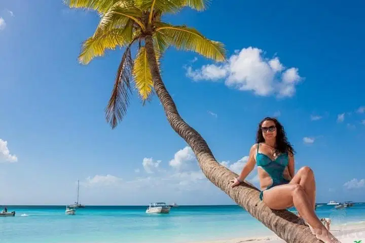 A woman in a teal bikini and sunglasses sits on a curved palm tree overhanging a sandy beach. The turquoise ocean, boats, and bright sky create the perfect scene to experience with the best tours Punta Cana has to offer.